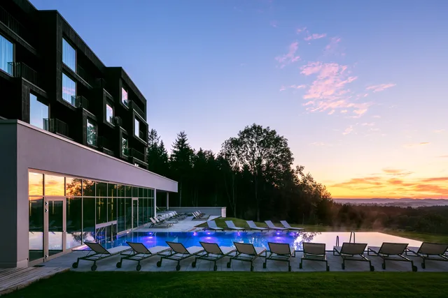Modern building with a glass facade and illuminated pool at sunset, surrounded by trees and lounge chairs.